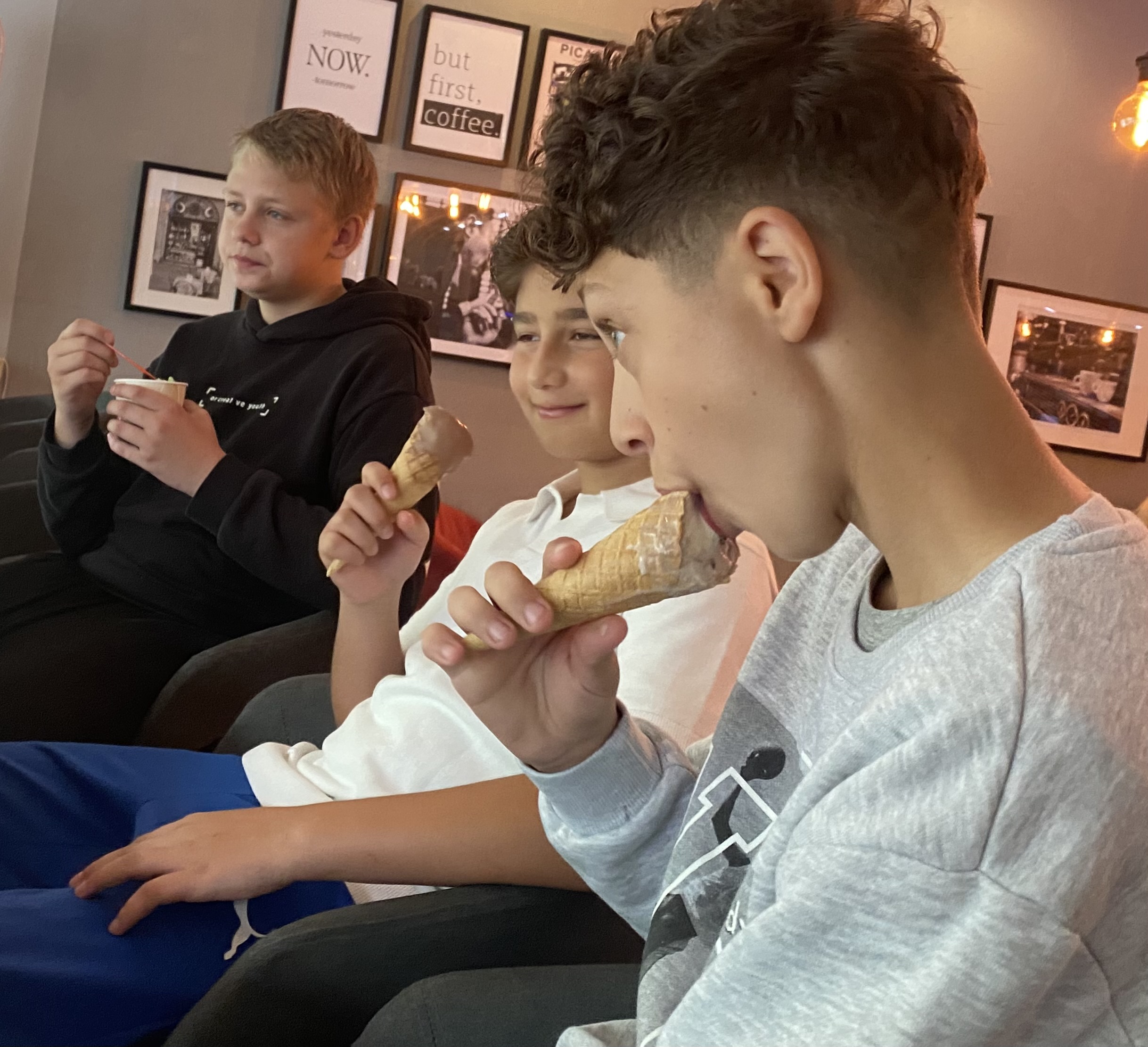 Drei Jungen sitzen nebeneinander in einem Café auf gepolsterten Stühlen und genießen ihr Eis. Zwei von ihnen essen Eiswaffeln, einer isst aus einem Becher mit Löffel. Im Hintergrund hängen stilvolle Bilderrahmen mit Sprüchen und Fotos an einer grauen Wand. Die Atmosphäre wirkt entspannt und fröhlich.