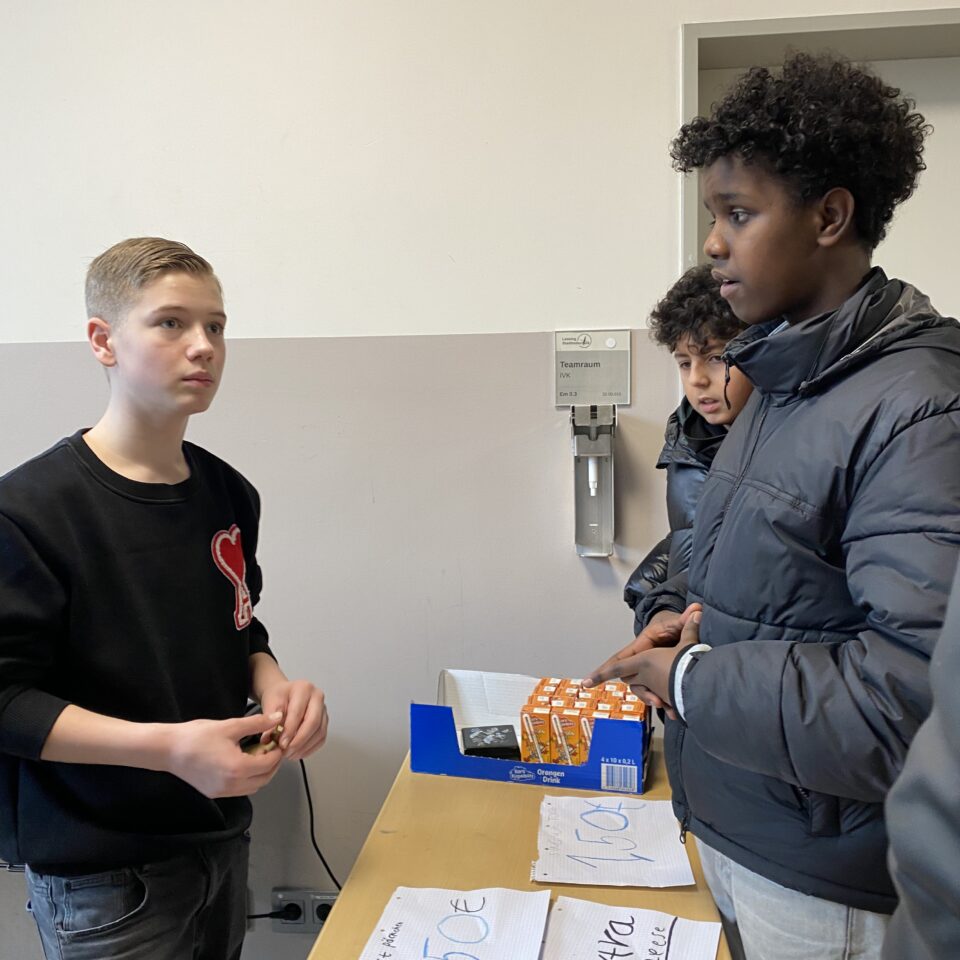 Schüler:innen stehen an einem Verkaufstisch mit Getränkepäckchen und handgeschriebenen Preisschildern. Eine Person hält Geld in den Händen.