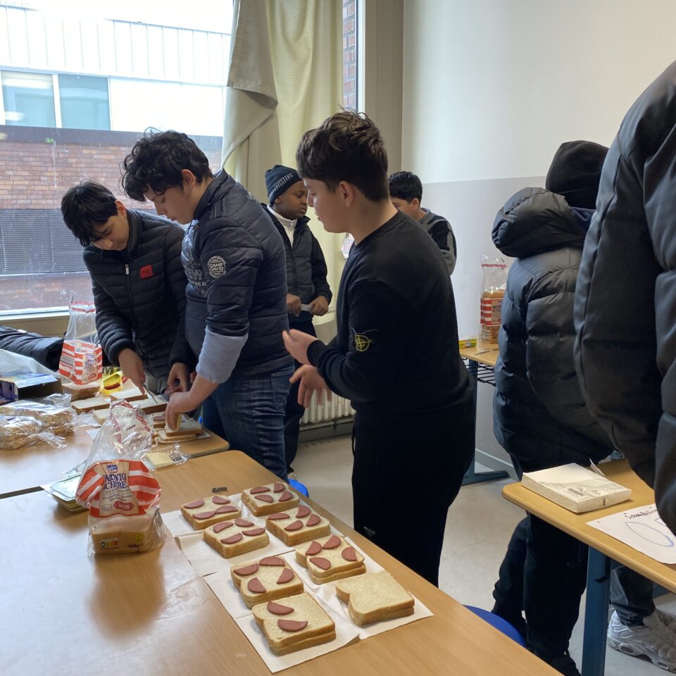 Schüler:innen der Klasse 7b bereiten gemeinsam Sandwiches vor. Auf den Tischen liegen Toastscheiben, Käse und Wurst.