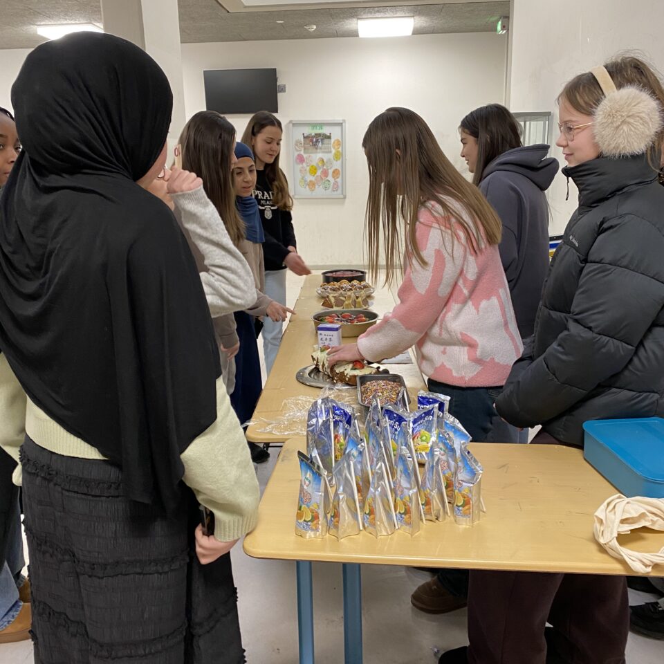 Schüler:innen bedienen am Kuchenstand der Klasse 7b ihre Mitschüler:innen. Auf den Tischen stehen verschiedene Kuchen und Getränkebeutel.