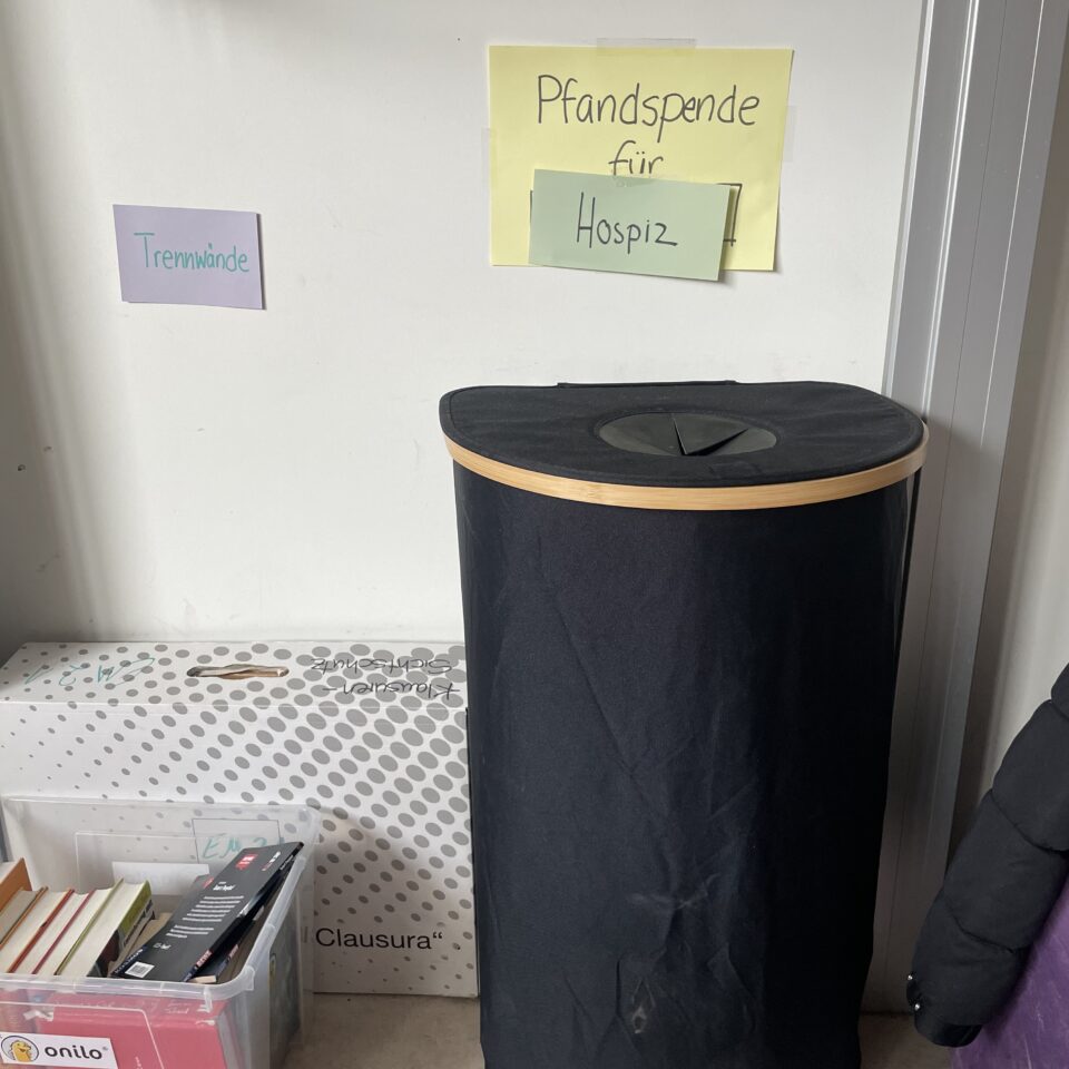 Schwarzer Sammelbehälter im Klassenraum der 8a. Darüber hängt ein handgeschriebenes Schild mit der Aufschrift „Pfandspende für Hospiz“.