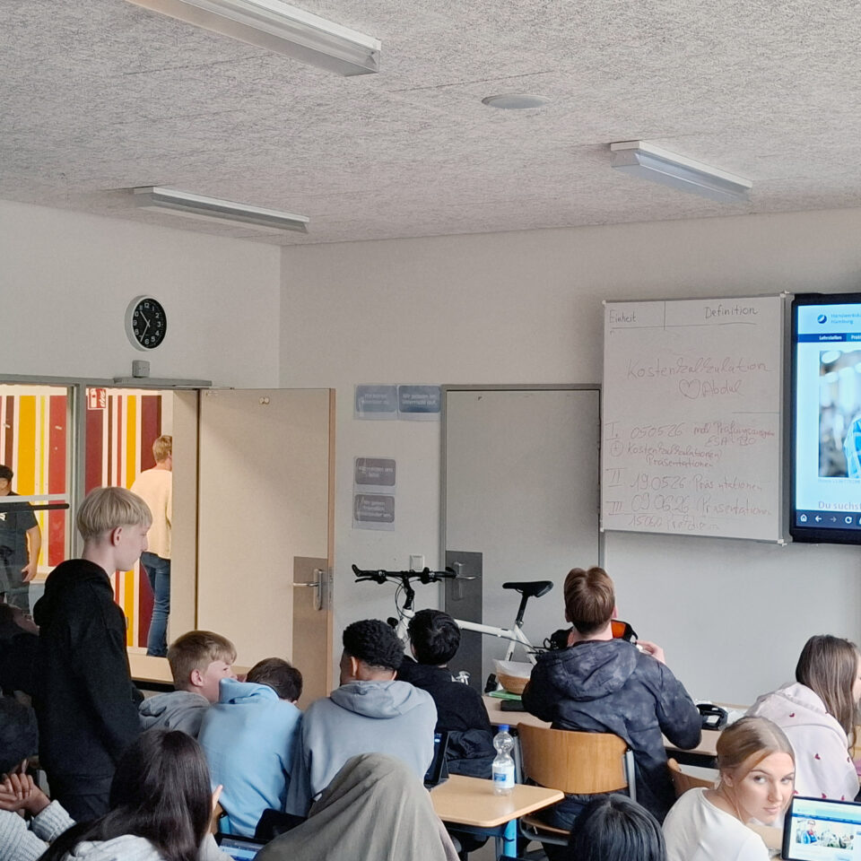 Schüler:innen sitzen und stehen in kleinen Gruppen im Klassenraum und arbeiten mit Tablets zur Berufsorientierung. Auf dem großen Bildschirm ist die Praktikumsbörse der Handwerkskammer Hamburg geöffnet.