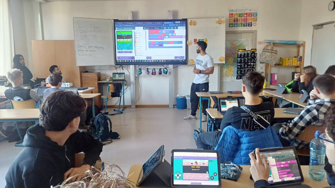 Schüler:innen der Lessing-Stadtteilschule programmieren beim Workshop der Hacker School eigene Spiele auf Tablets. Auf den Geräten und dem großen Bildschirm sind farbige Programmierblöcke und Spielansichten zu sehen.