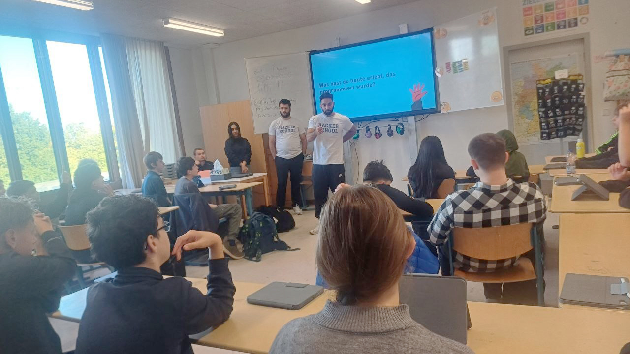 Zwei Mitarbeiter der Hacker School stehen vor der Klasse und besprechen mit den Schüler:innen Beispiele für Programmierung im Alltag. Auf dem Bildschirm steht die Frage: „Was hast du heute erlebt, das programmiert wurde?“