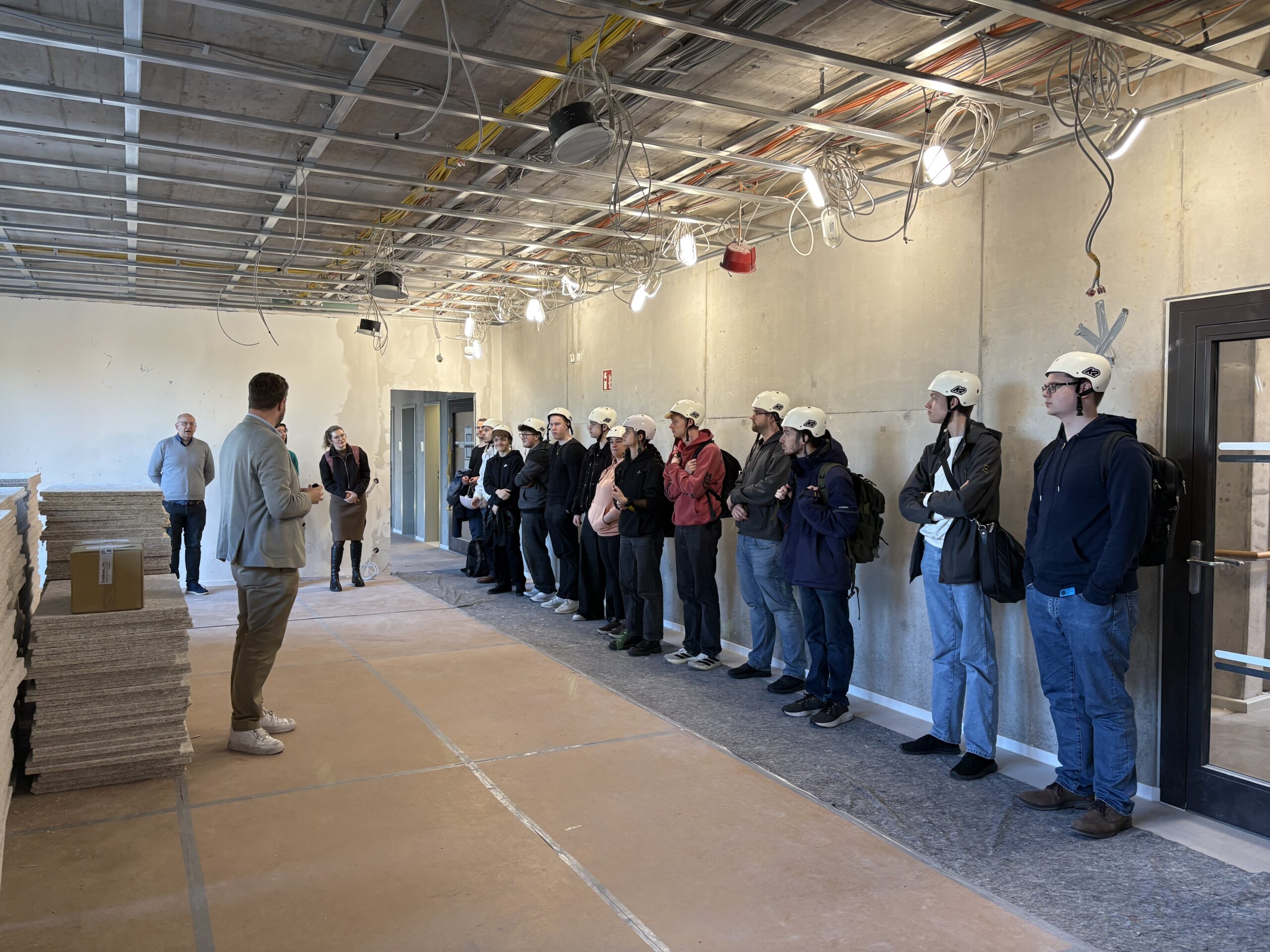 Herr Hartten spricht zur Besuchergruppe im künftigen CoLab. Die Schülerinnen und Schüler stehen mit weißen Helmen nebeneinander an der Wand und hören zu.