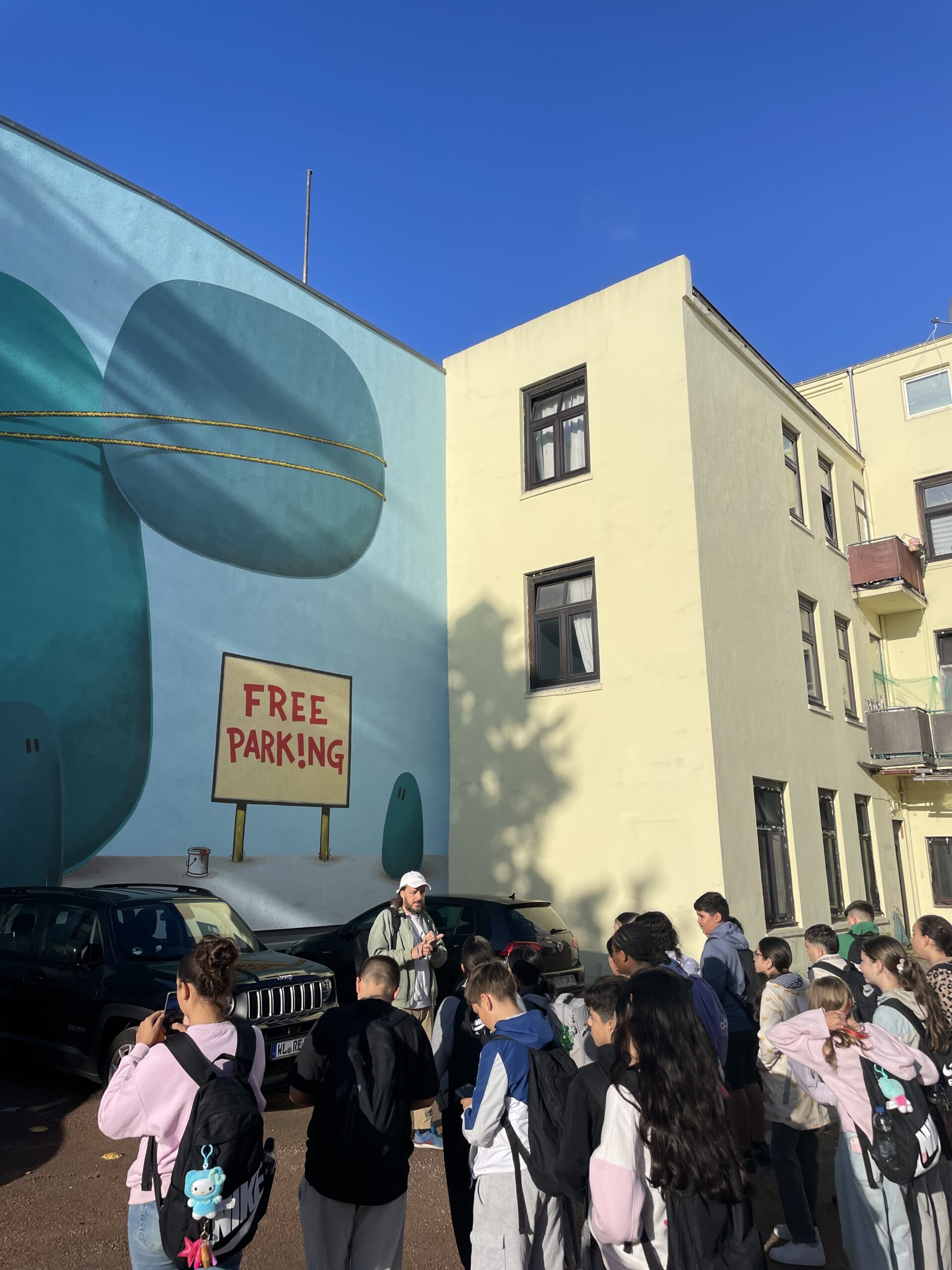 Die Gruppe steht vor einem großflächigen Wandbild mit türkisfarbenen Fantasiefiguren und dem Schriftzug „Free Parking“.