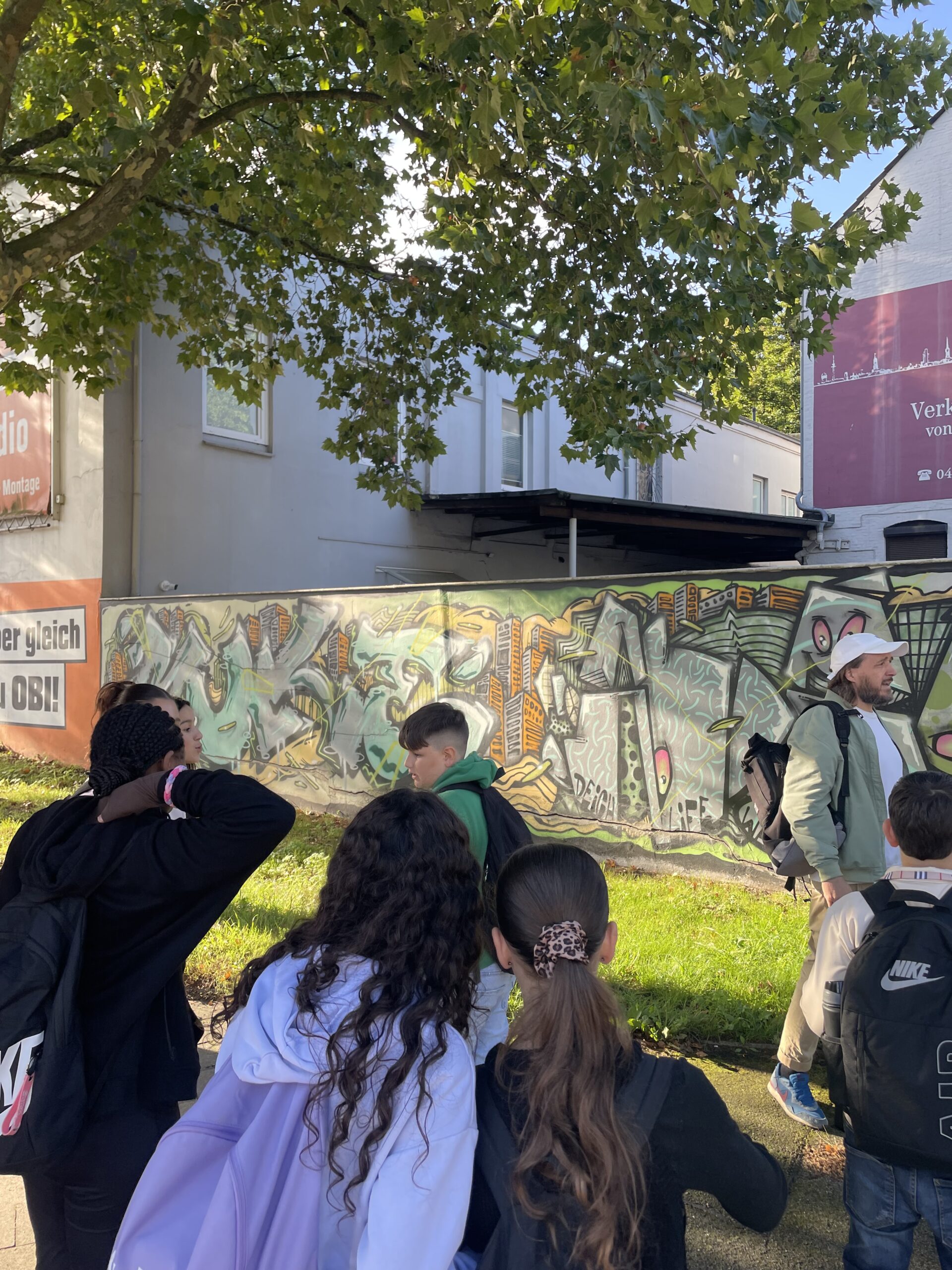 Schülerinnen und Schüler und Stadtführer vor einer Graffitiwand mit grün-grauen Schriftzügen und orangefarbenen Hochhäusern.