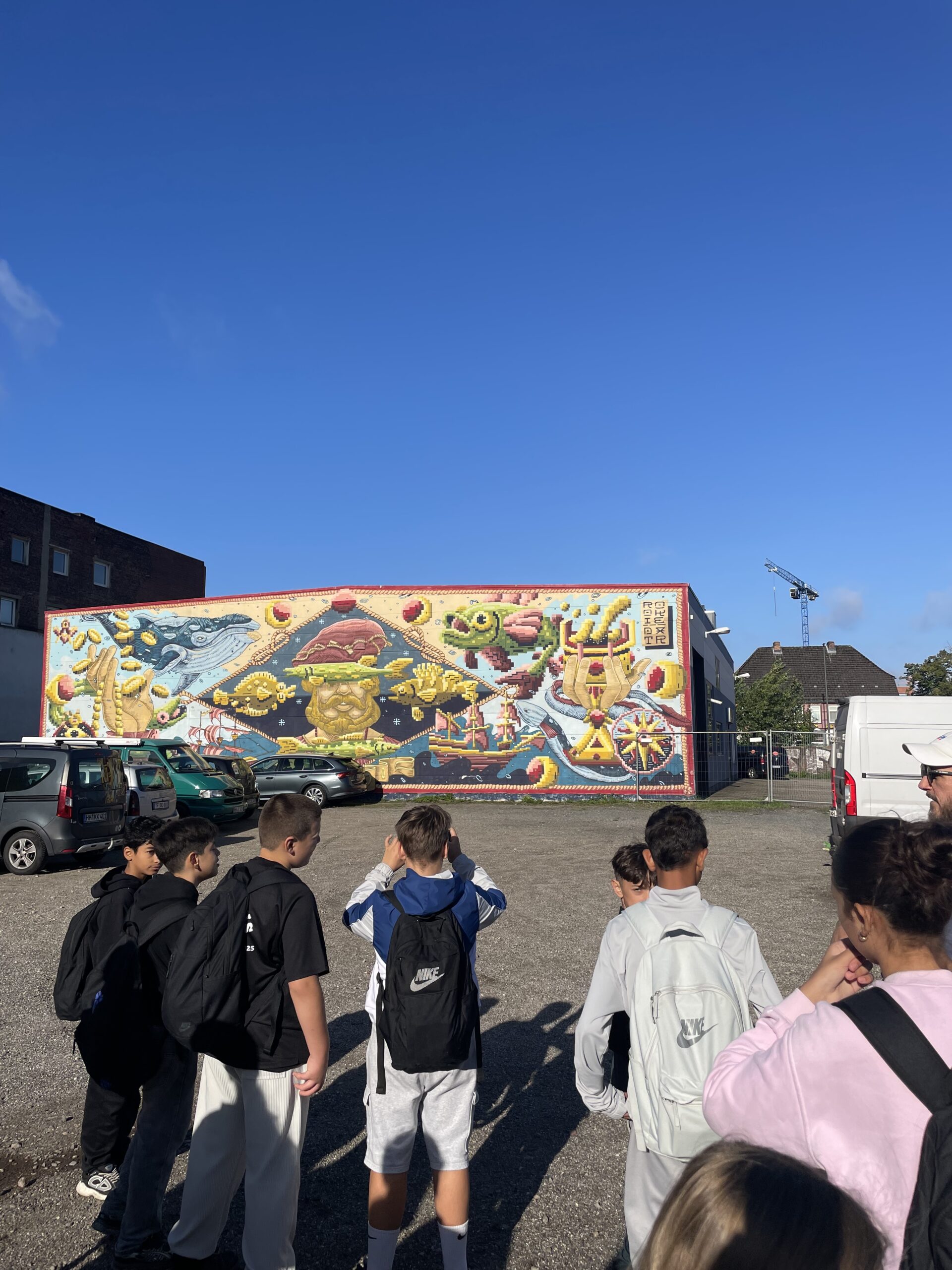 Schülerinnen und Schüler vor einem bunten Wandbild im Pixelstil mit einem bärtigen Mann, Walen, Segelschiffen und Goldmünzen.