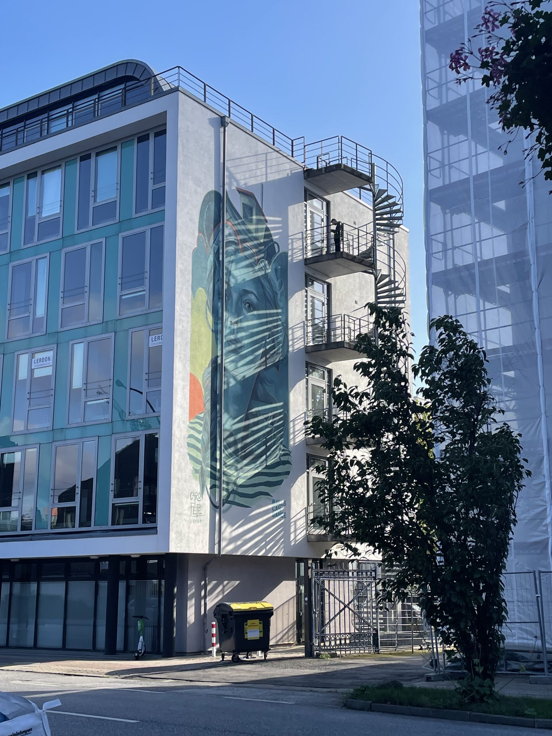 Mehrere Stockwerke hohes Wandbild eines Gesichts in Blau- und Grüntönen. Eine benachbarte Wendeltreppe wirft Schattenstreifen auf das Motiv.